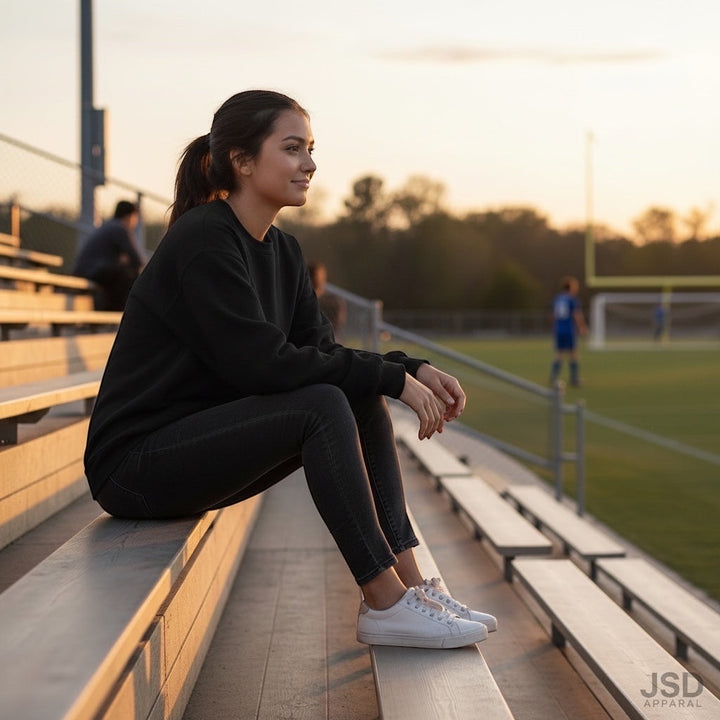 A woman wearing a JSD Apparel black fleece crewneck sweatshirt sitting on stadium bleachers at sunset.