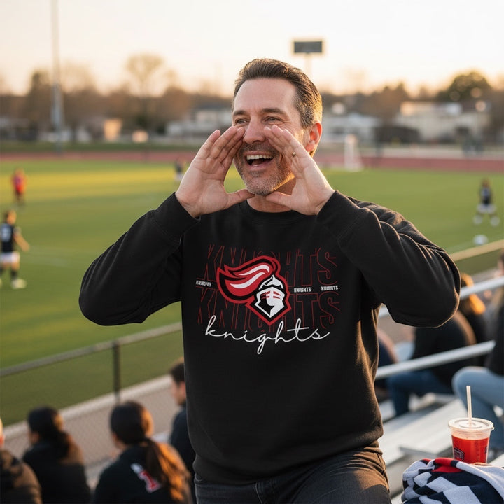 JSD Apparel black crewneck sweatshirt with Knights script graphic worn by a man cheering at a sports field.