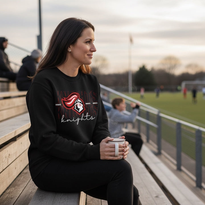 JSD Apparel black crewneck sweatshirt with Knights collegiate graphic, worn by a woman sitting on stadium bleachers.