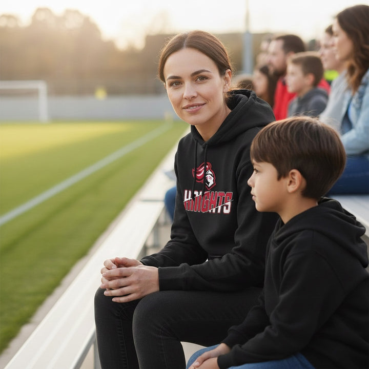 JSD Apparel black heavyweight hoodie with Hillcrest Knights graphic worn by a woman sitting at a sports field