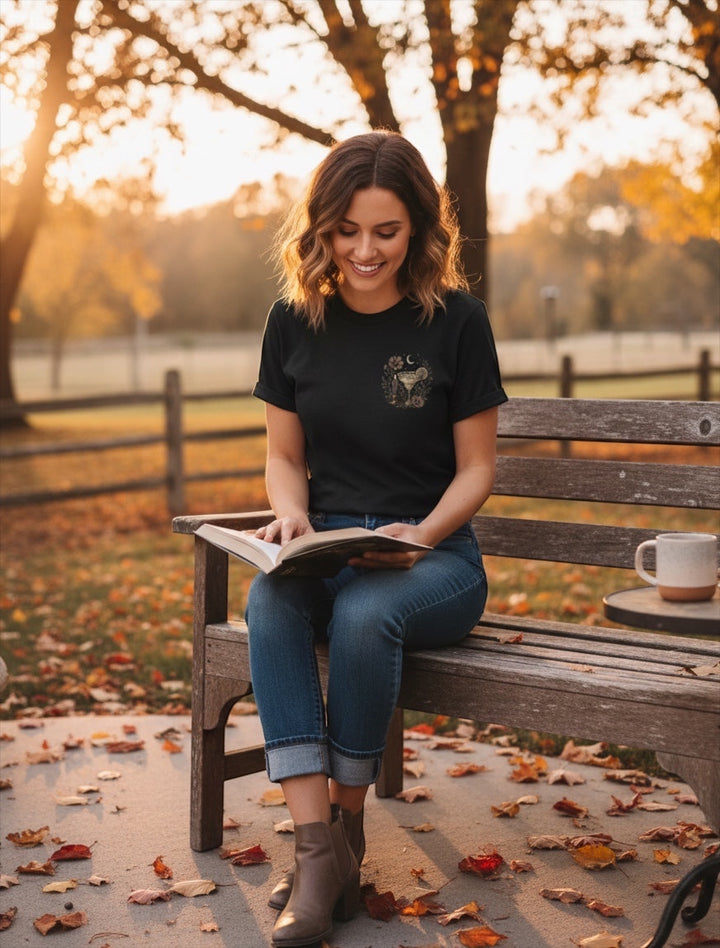 JSD Apparel black unisex tee with a small margarita chest graphic, worn by a woman reading on a park bench in autumn.