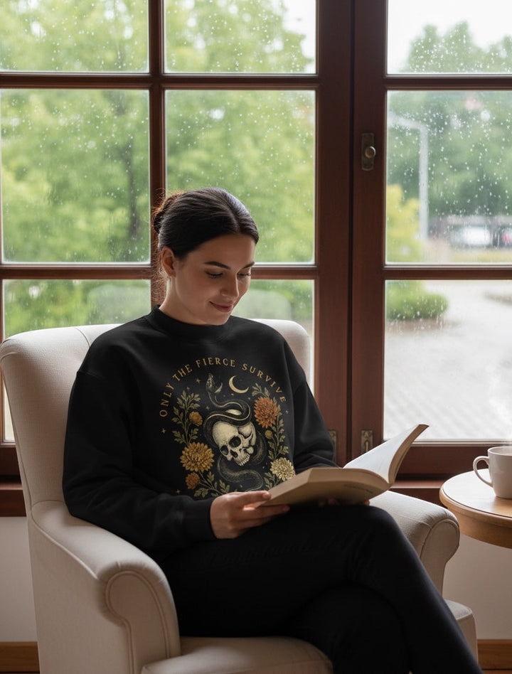 JSD Apparel black mock neck sweatshirt with skull, serpent, and floral graphic worn by a woman reading in a chair.