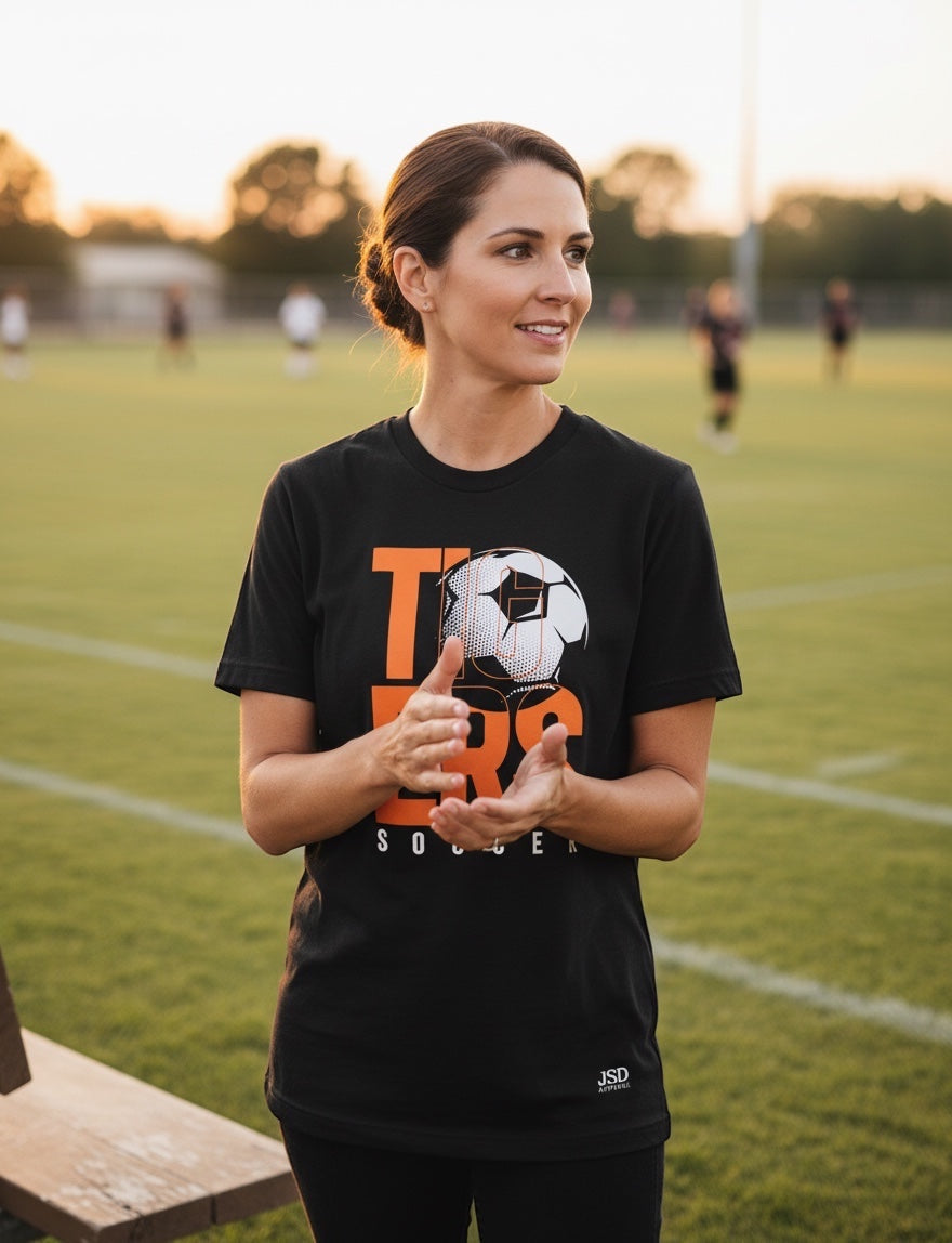 JSD Apparel black unisex jersey tee with orange and white TIGERS soccer graphic worn by a woman on a sports field.