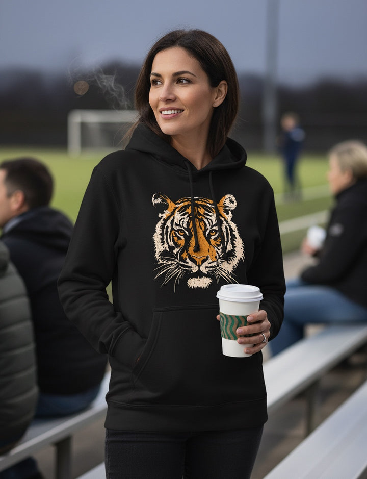 Woman wearing a black JSD Apparel heavyweight hoodie with a vintage tiger graphic, holding a coffee at a sports field.