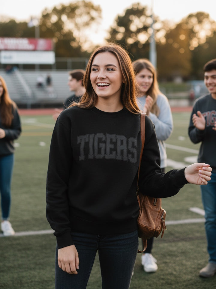JSD Apparel black crewneck sweatshirt with TIGERS arch lettering worn by a smiling woman on a sports field