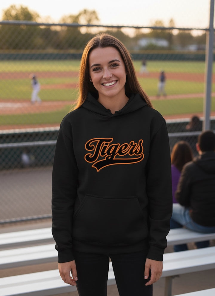 A smiling woman wearing a black JSD Apparel heavyweight hoodie with an orange Tigers script graphic at a baseball field.