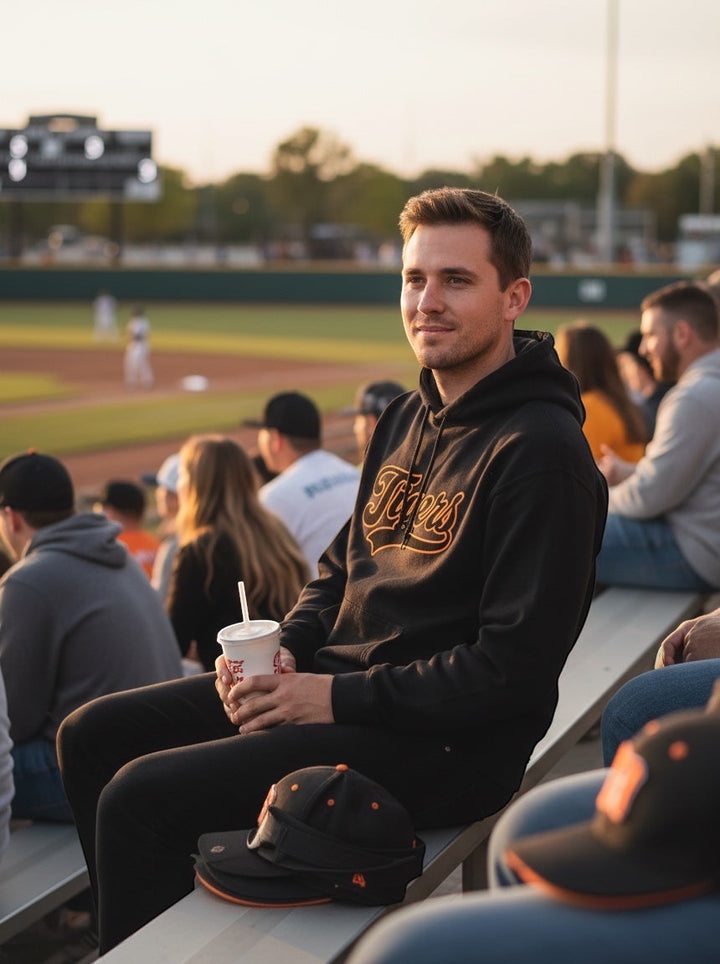 Man wearing a JSD Apparel black heavyweight hoodie with orange Tigers script at a baseball game.