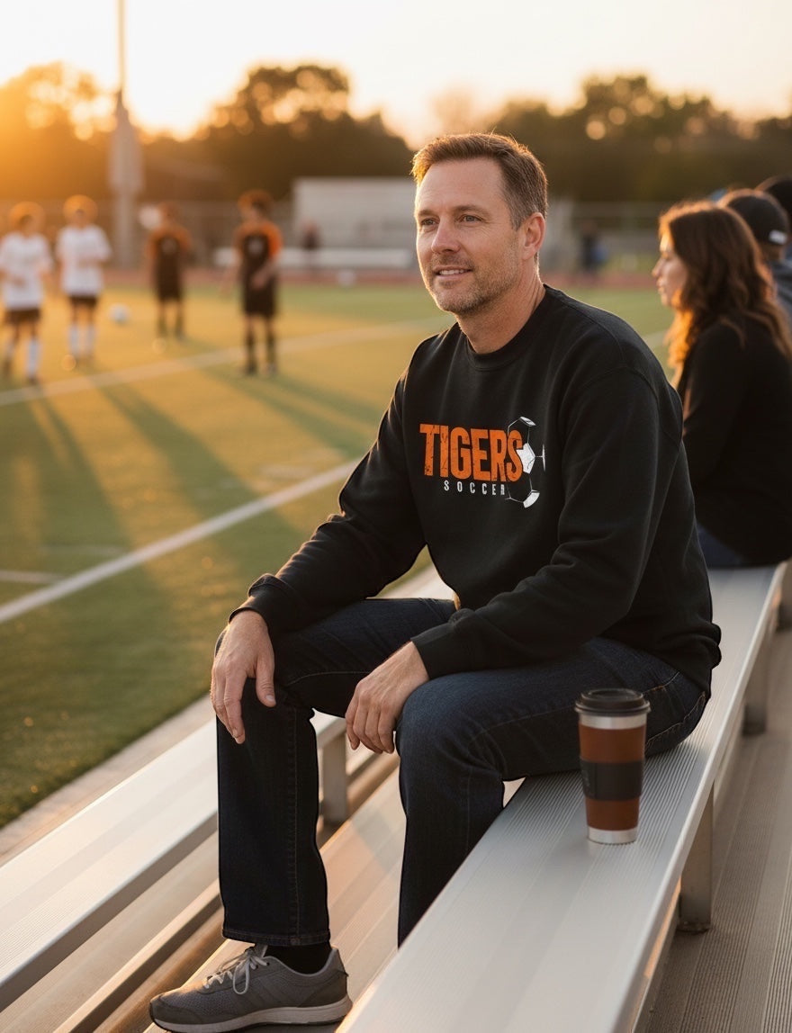 JSD Apparel black vintage crewneck sweatshirt with Tigers Soccer graphic worn by a man sitting on a soccer field bench