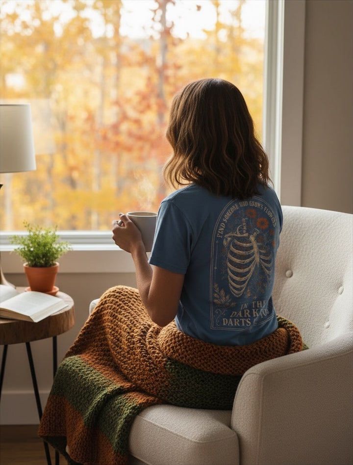 JSD Apparel blue Comfort Colors unisex tee with skeletal floral back graphic, worn by a woman sitting in a chair.