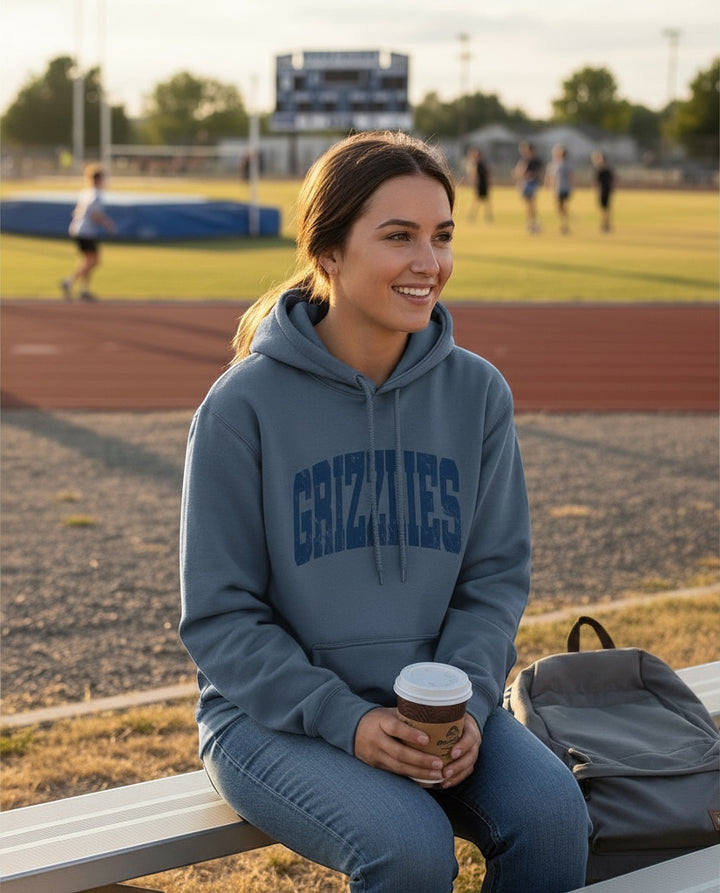 A woman wearing a blue JSD Apparel Skyline Grizzlies Arch Hoodie with a distressed collegiate print on a sports field.