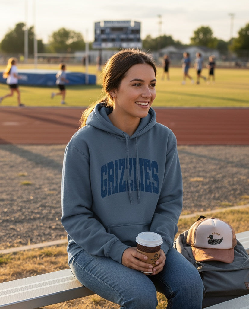 Woman wearing a blue JSD Apparel Skyline Grizzlies arch hoodie with a distressed collegiate print on a sports field.