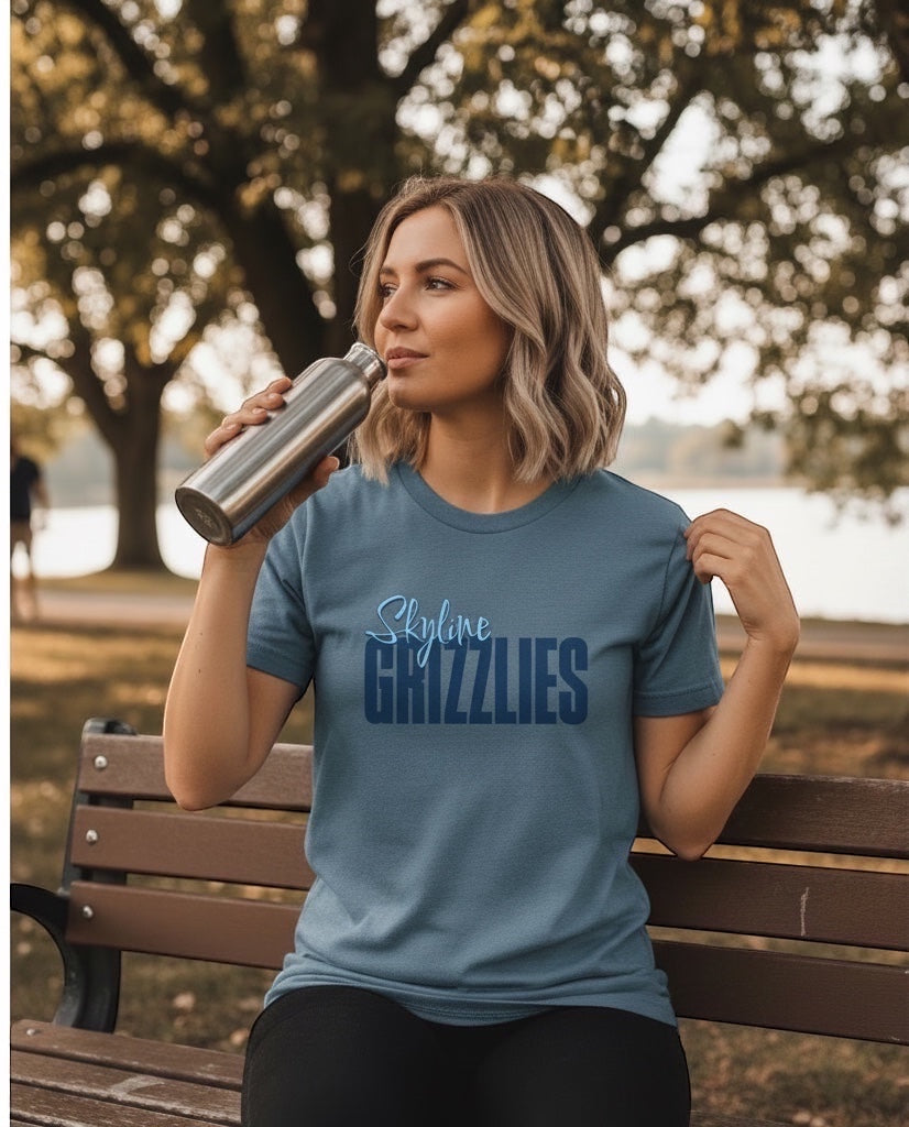 Woman wearing a blue JSD Apparel Skyline Grizzlies script cotton tee while drinking from a metal bottle in a park.
