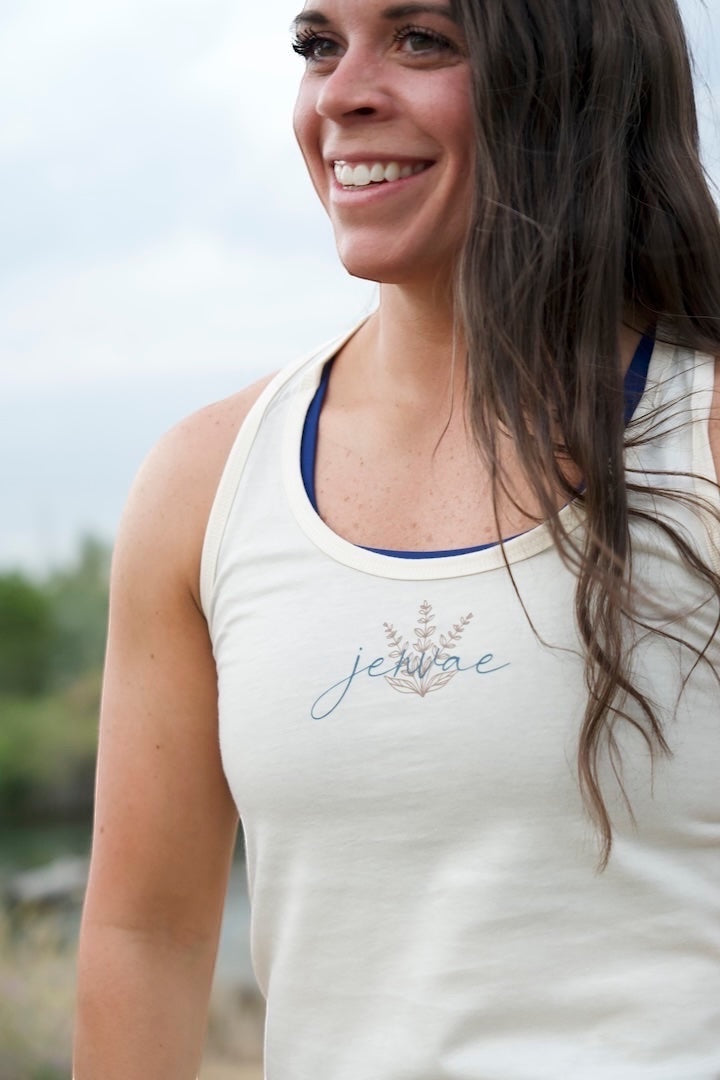 JSD Apparel bone white cotton blend racerback tank with Jenvae botanical logo design. Model is wearing a blue sports bra underneath.