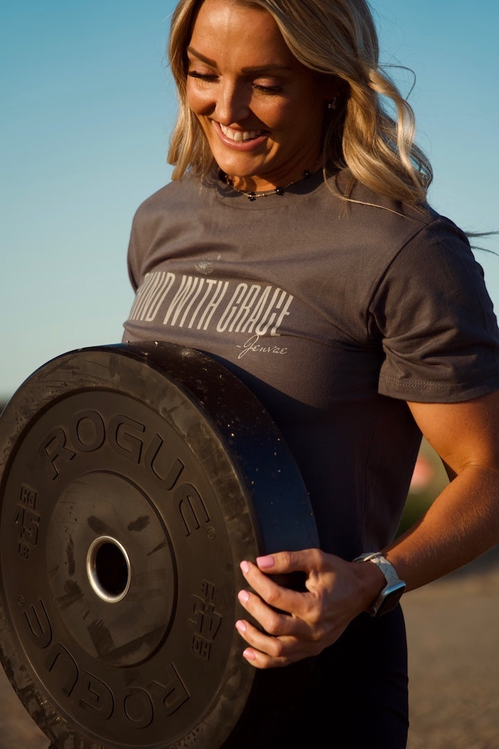 JSD Apparel charcoal cropped tee with Grind With Grace graphic, worn by a smiling woman holding a weight plate outdoors.
