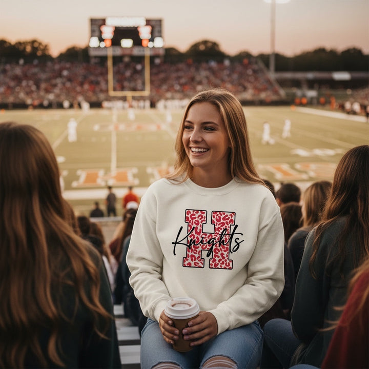 JSD Apparel cream heavyweight crewneck sweatshirt with a red leopard-print H and black Knights script at a stadium.