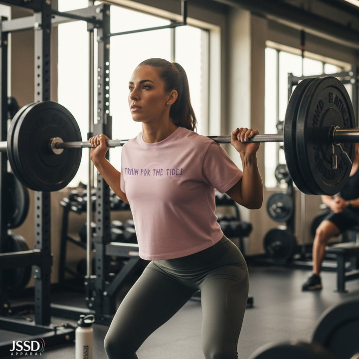 JSD Apparel light pink cropped tee with "Train for the Tides" graphic, worn by a woman weightlifting.