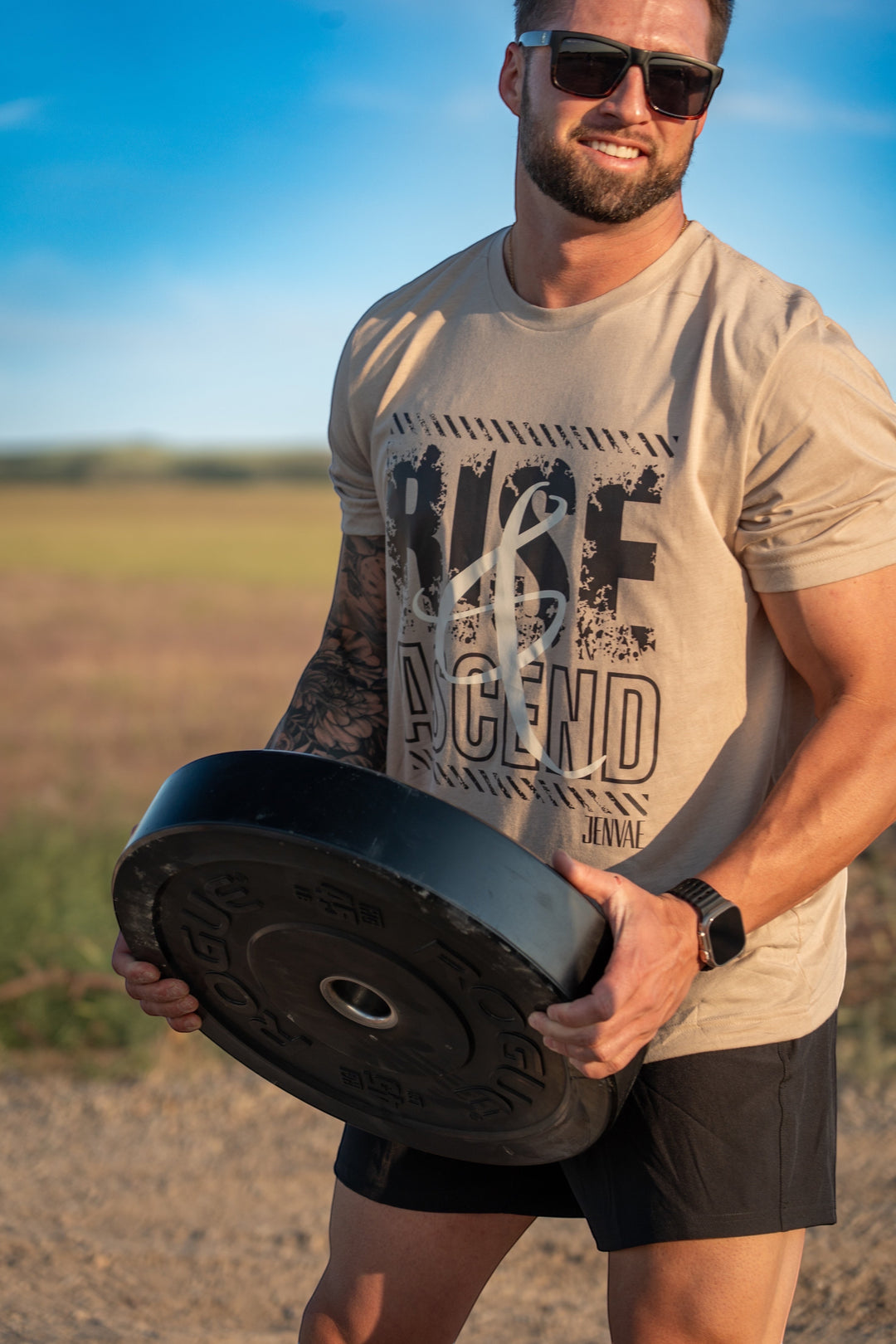 JSD Apparel desert sand unisex cotton t-shirt with distressed Rise & Ascend graphic worn by a man holding a weight plate.