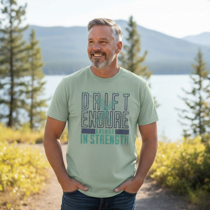 Man wearing a sage green JSD Apparel cotton t-shirt with Drift & Endure graphic, standing by a lake in nature.