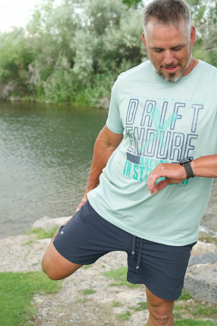 Man wearing a light green JSD Apparel Drift & Endure cotton t-shirt while stretching by a river.