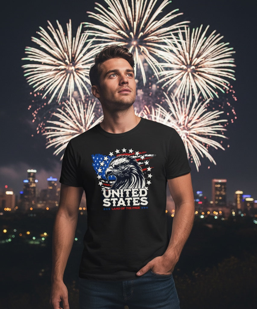 Man wearing JSD Apparel black patriotic t-shirt with eagle and flag graphic against a fireworks city skyline background