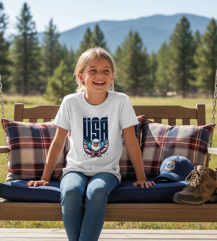 A smiling girl wearing a JSD Apparel white youth tee with a patriotic Freedom Eagle USA graphic on a wooden swing.