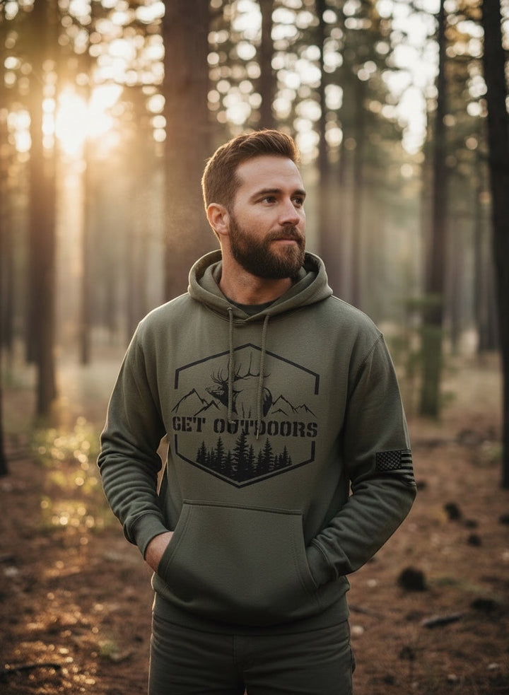 Man wearing JSD Apparel olive green Get Outdoors Elk unisex fleece hoodie with graphic print in a forest setting.