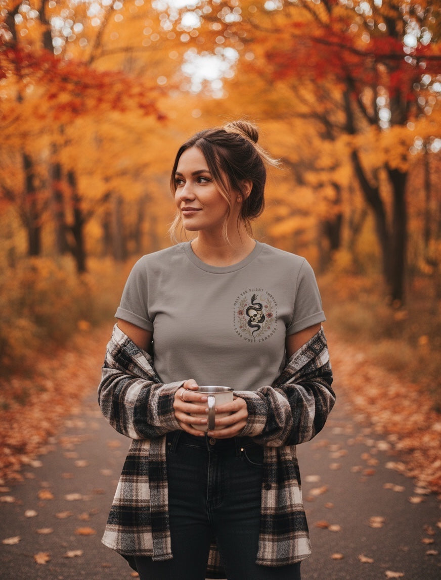 JSD Apparel unisex tee in grey with a small chest graphic of a skull and serpent, worn by a woman in a fall forest.