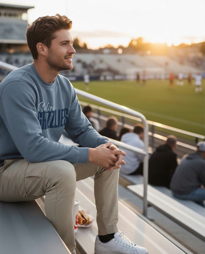 Man wearing a blue JSD Apparel Skyline Grizzlies crewneck sweatshirt while sitting in stadium bleachers at sunset.