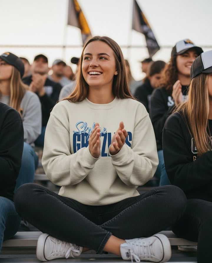 A woman wearing a JSD Apparel Skyline Grizzlies crewneck sweatshirt, sitting in stadium bleachers and clapping.