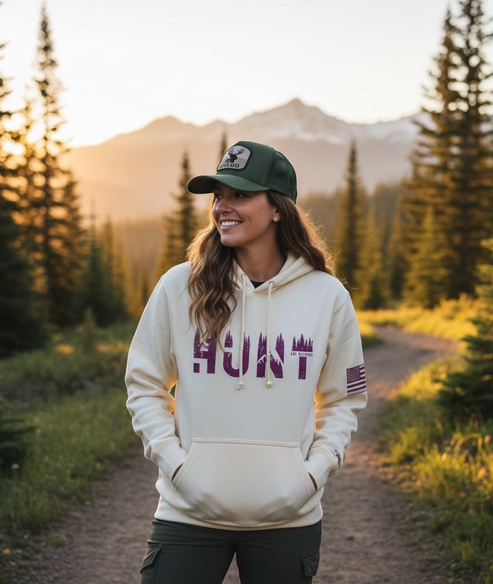 JSD Apparel unisex cream pullover hoodie with HUNT graphic, worn by a woman in a mountain forest at sunset.