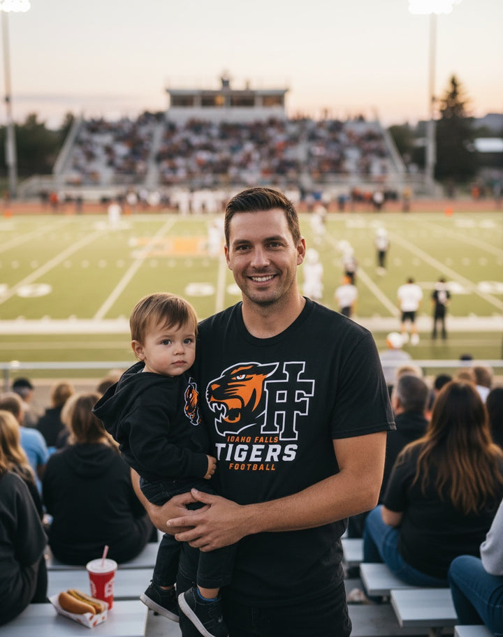 JSD Apparel black Idaho Falls Tigers football crewneck tee worn by a man holding a toddler at a stadium game