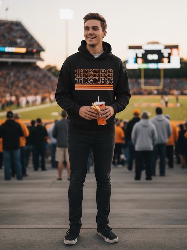 Man wearing a black JSD Apparel Idaho Falls Tigers retro stripe hoodie with orange graphic at a football stadium.