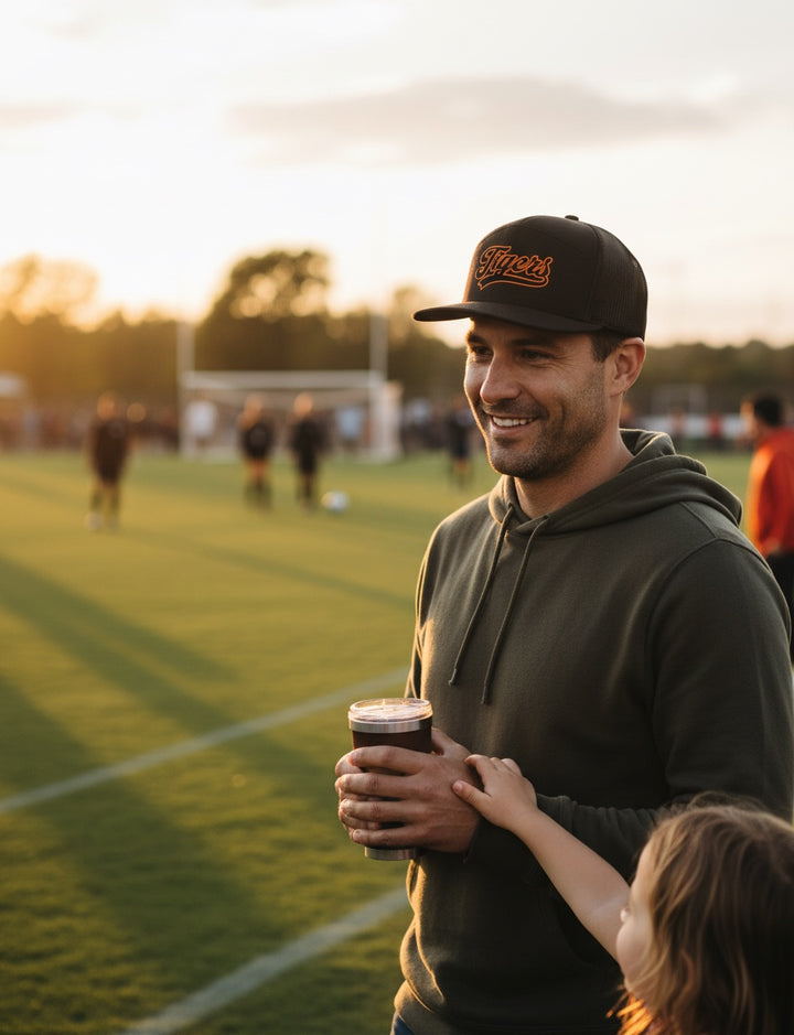 JSD Apparel Idaho Falls Tigers black performance hat with orange script logo worn by a man at a soccer field