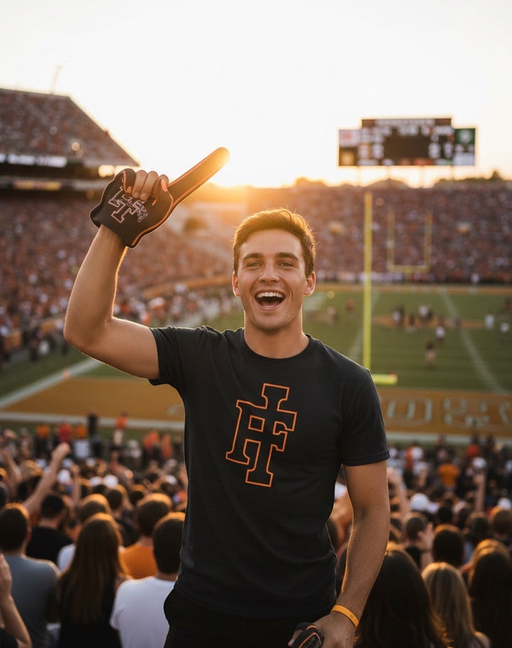 JSD Apparel Idaho Falls Tigers black premium tee with orange IF logo worn by a smiling fan at a football stadium