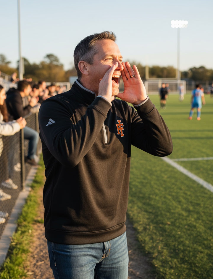 JSD Apparel Idaho Falls Tigers black Adidas quarter-zip pullover worn by a man cheering on a sports field