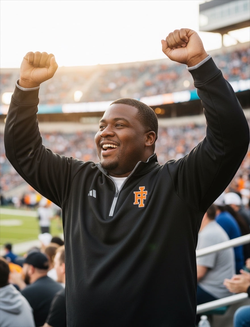 JSD Apparel Idaho Falls Tigers black Adidas quarter-zip pullover worn by a cheering fan at a stadium.