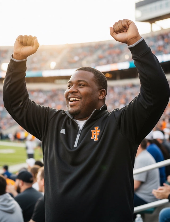 JSD Apparel Idaho Falls Tigers black Adidas quarter-zip pullover worn by a cheering fan at a stadium.