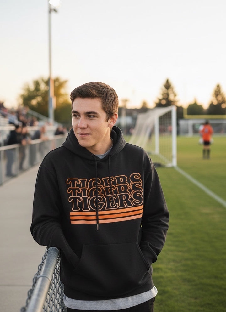 Man wearing a black JSD Apparel Idaho Falls Tigers retro hoodie with orange stacked text and stripes at a sports field