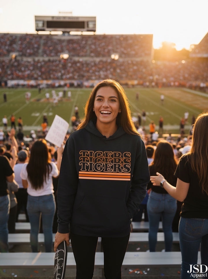 Woman wearing a JSD Apparel Idaho Falls Tigers navy hoodie with retro orange stripes at a sunset football game.