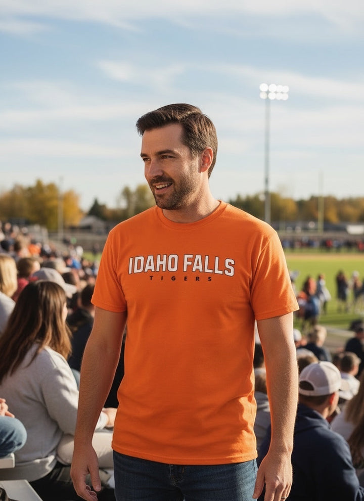 Man wearing an orange JSD Apparel Idaho Falls Tigers cotton t-shirt at a sunny outdoor sports stadium.
