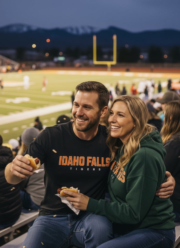 Man wearing a black JSD Apparel Idaho Falls Tigers cotton tee at a football game with a woman in a green hoodie.