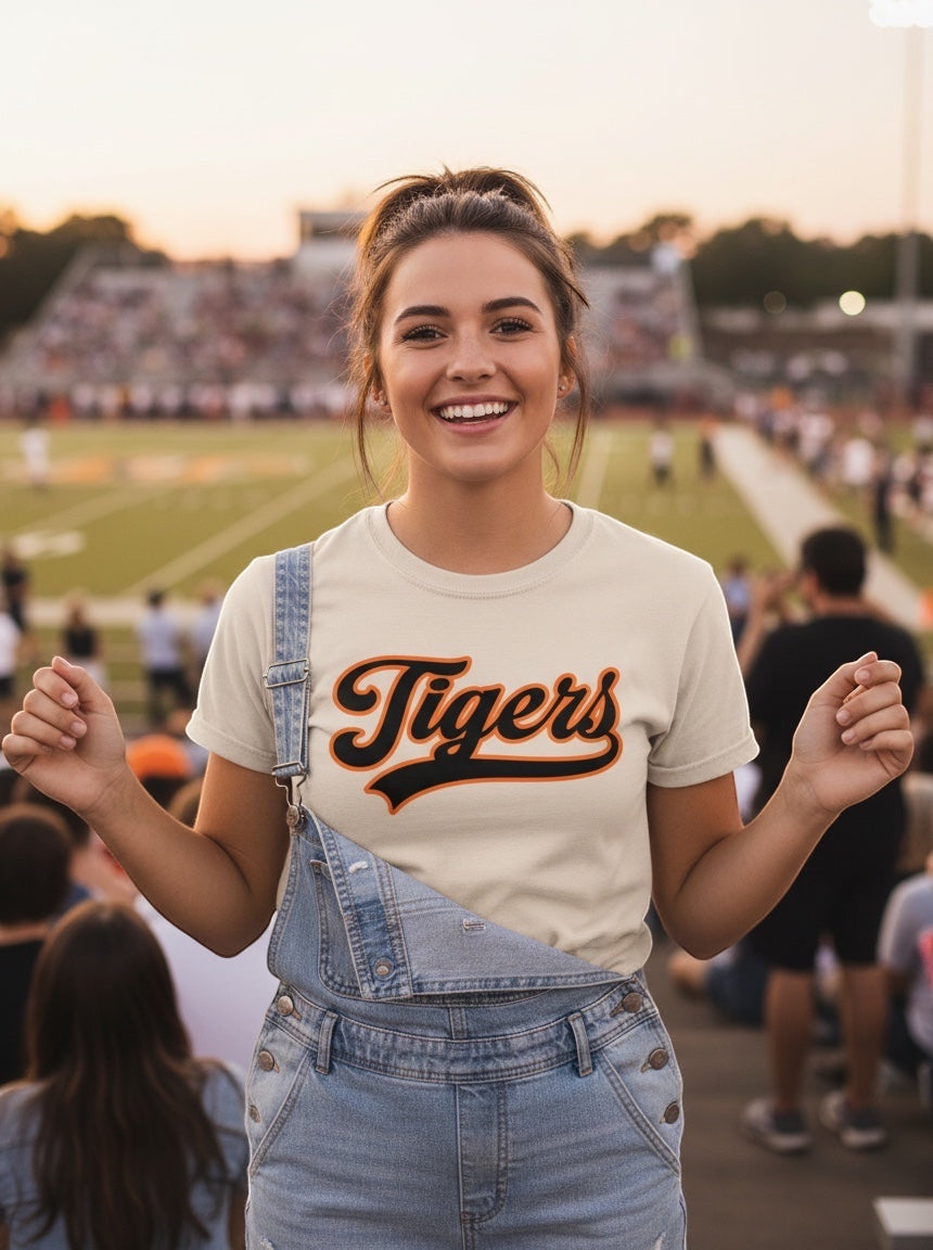A woman wearing a JSD Apparel Idaho Falls Tigers cream cotton tee with orange and black script at a football game.