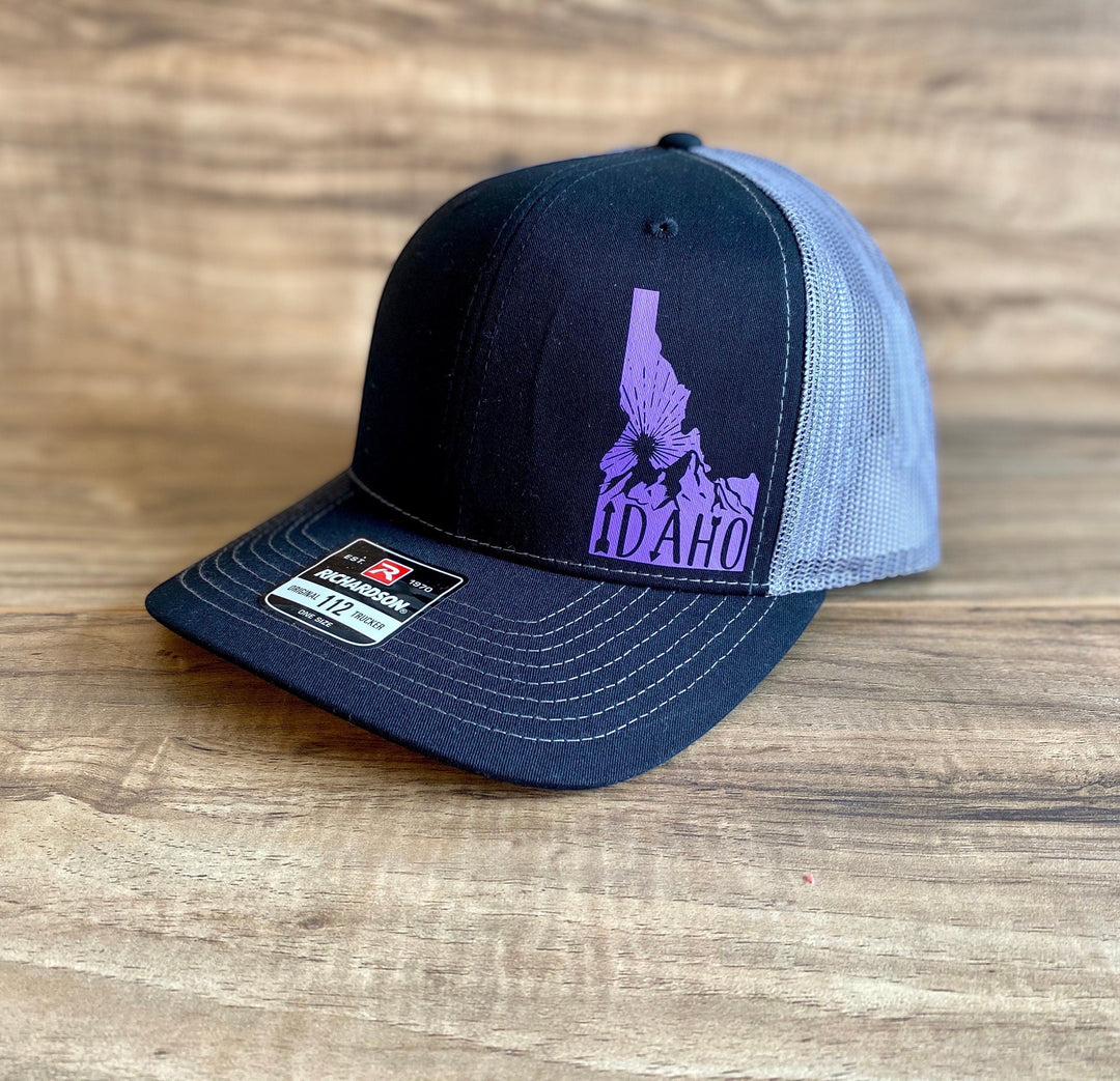JSD Apparel Idaho Sunrise Richardson 115 trucker hat with black front, grey mesh back, and purple Idaho graphic.
