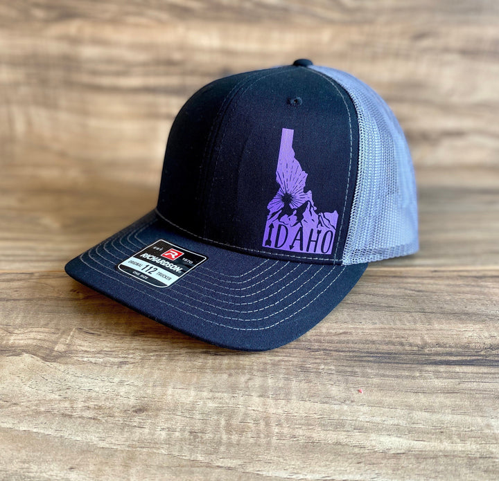 JSD Apparel Idaho Sunrise Richardson 115 trucker hat with black front, grey mesh back, and purple Idaho graphic.
