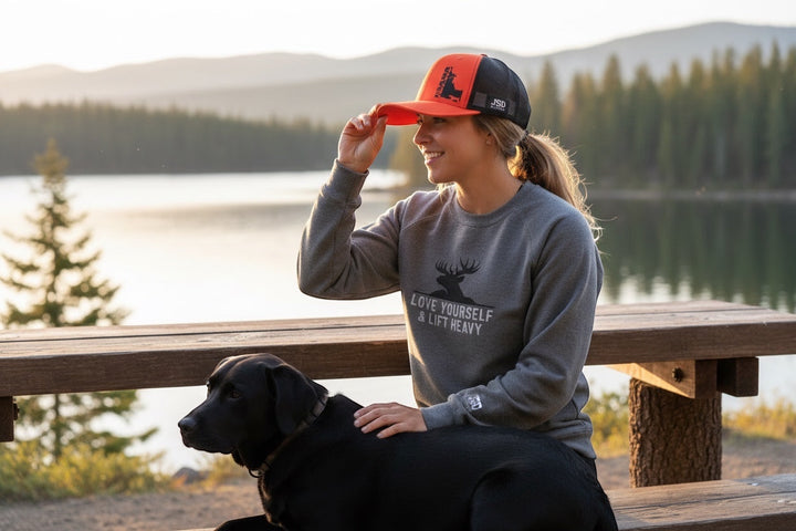 JSD Apparel Idaho Upland Blaze trucker hat with orange front and black mesh back, worn by a woman with a black dog.