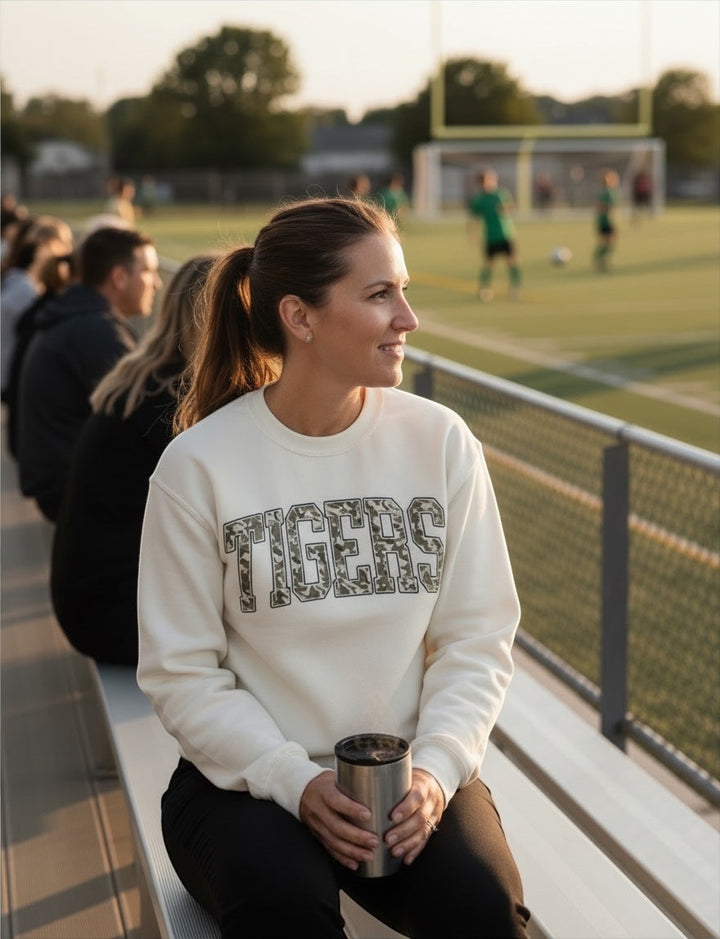 Woman wearing an ivory JSD Apparel Tigers crewneck sweatshirt with camo lettering while sitting at a sports field.