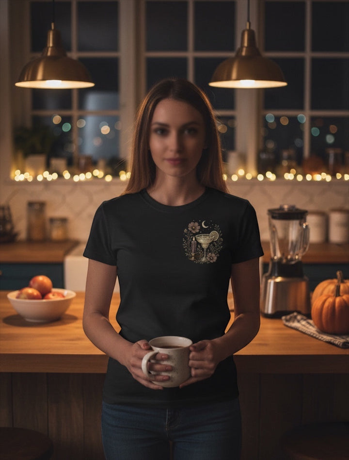 A woman wearing a JSD Apparel black unisex tee with a small margarita graphic on the chest in a cozy kitchen setting.