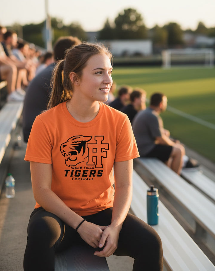 JSD Apparel orange Idaho Falls Tigers football crewneck t-shirt worn by a woman sitting on stadium bleachers