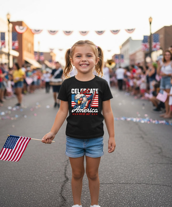 A young girl wearing a black JSD Apparel patriotic eagle graphic tee and denim shorts, holding a small American flag.