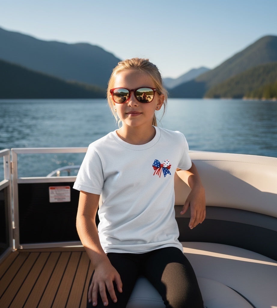 JSD Apparel youth white t-shirt with patriotic bow graphic, worn by a girl on a boat during a summer day.